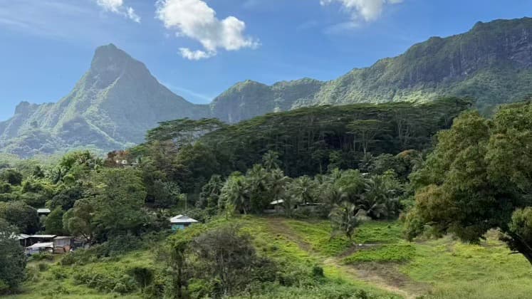 Lush green tropical valley with traditional houses nestled beneath towering volcanic mountain peaks.